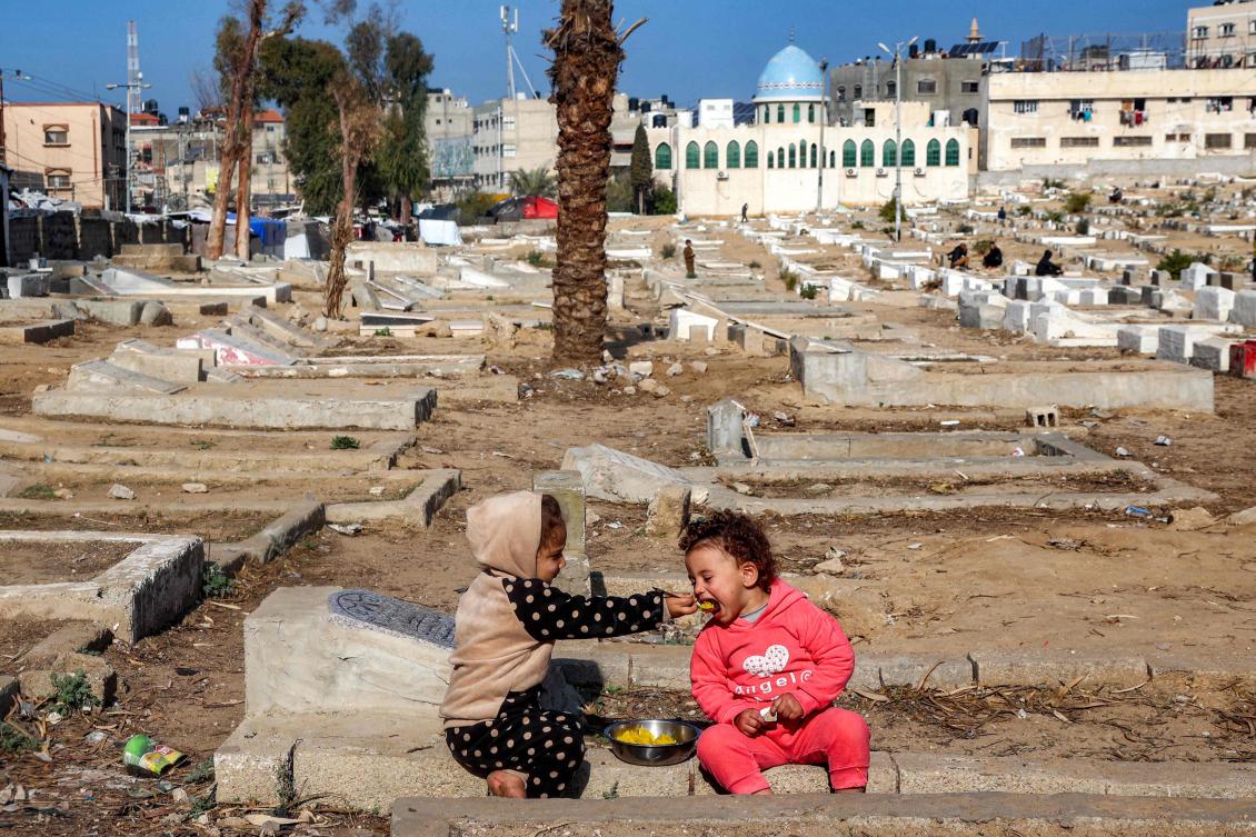 A child feeds another a spoonful of food as they sit atop graves at a cemetery where families displaced by conflict are taking shelter in Deir El-Balah in the central Gaza Strip on January 12, 2025. (AFP)
