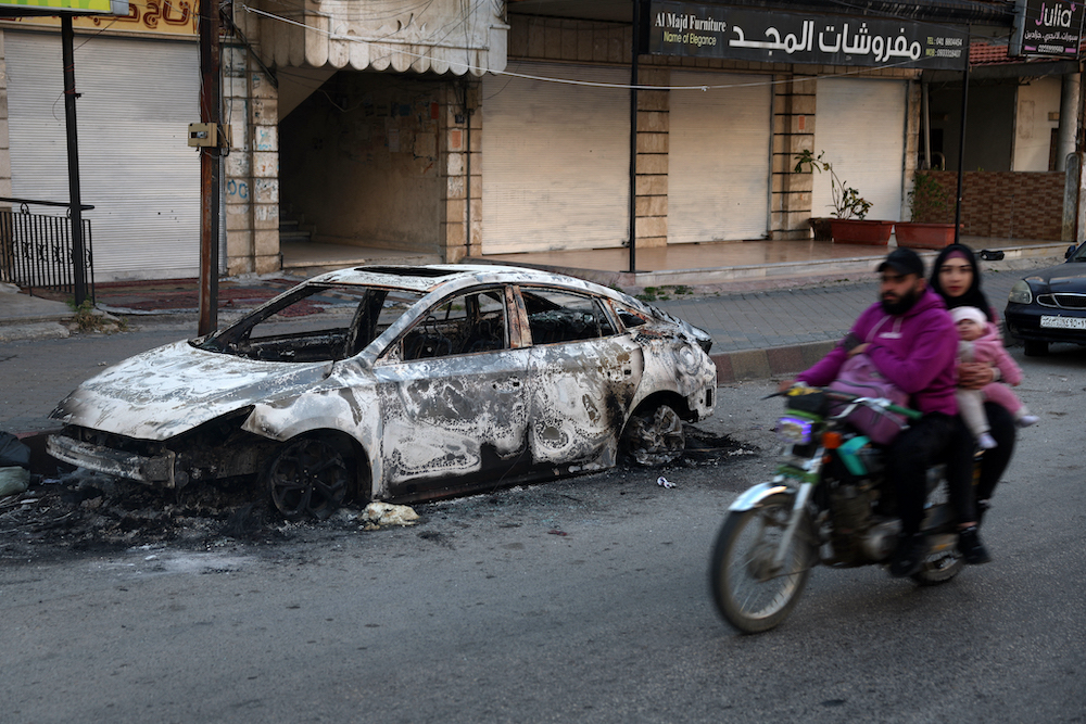 Members of a family riding a motorcycle drive past a charred car in the town of Jableh in Syria's coastal province of Latakia, Mar. 12, 2025. (AFP)