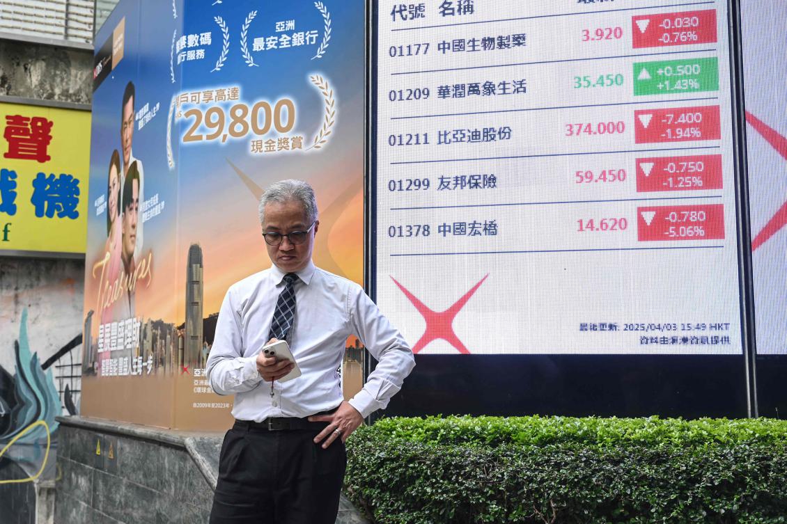 A man checks his phone next to an electronic board showing stocks on the Heng Seng Index in Hong Kong on Apr. 3, 2025. (AFP)
