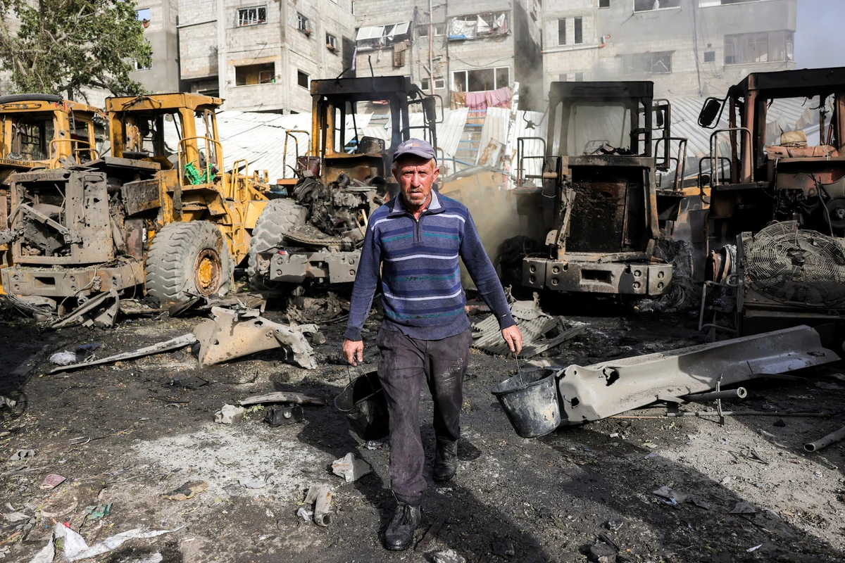 A man walks with empty pails of water in his hands past destroyed and burnt-down excavators at the Jabalia municipality garage in the northern Gaza Strip, Apr. 22, 2025. (AFP)