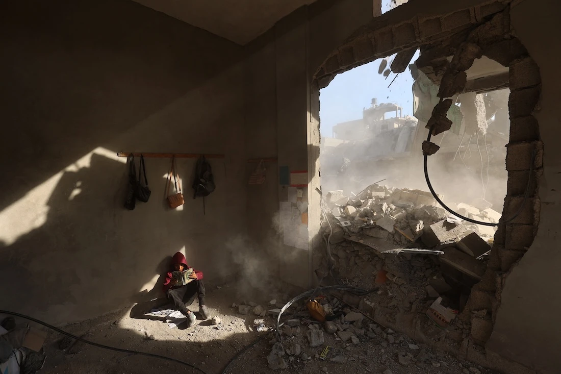 A Palestinian boy holds a book as he sits in rubble of a house, following overnight Israeli strikes. (File/AFP)