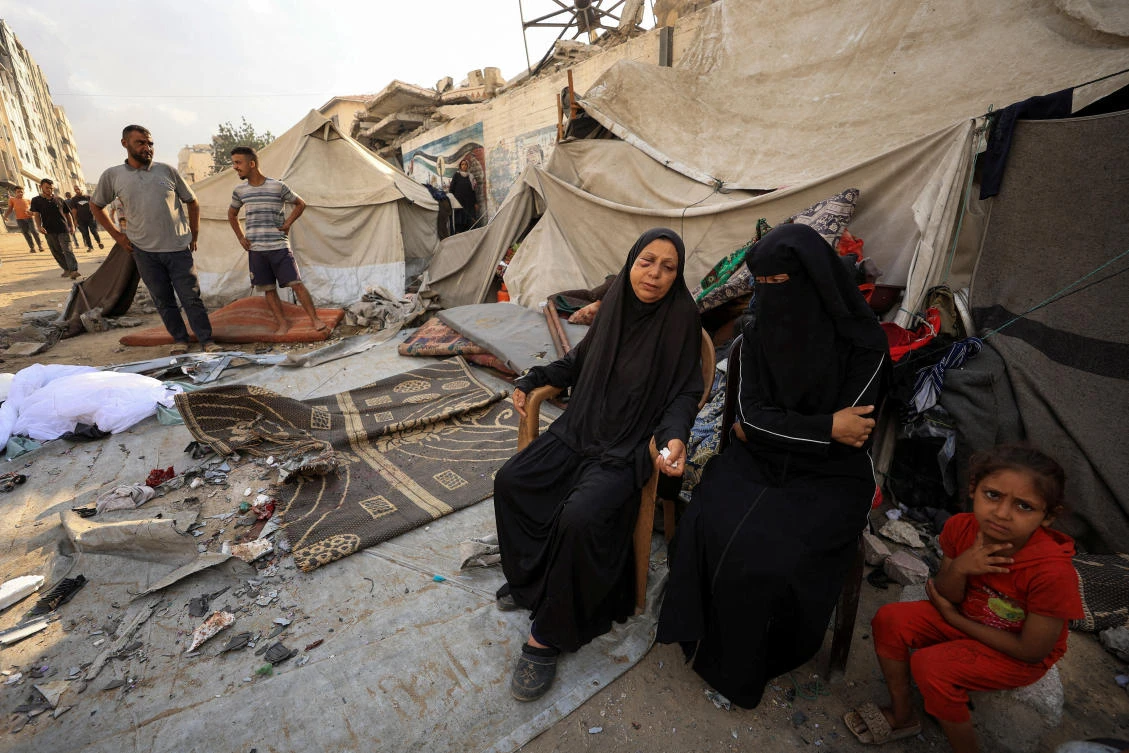 Palestinian women and a girl sit while others inspect the site of an overnight Israeli strike on a tent, in Gaza City, September 7, 2025. (Reuters)