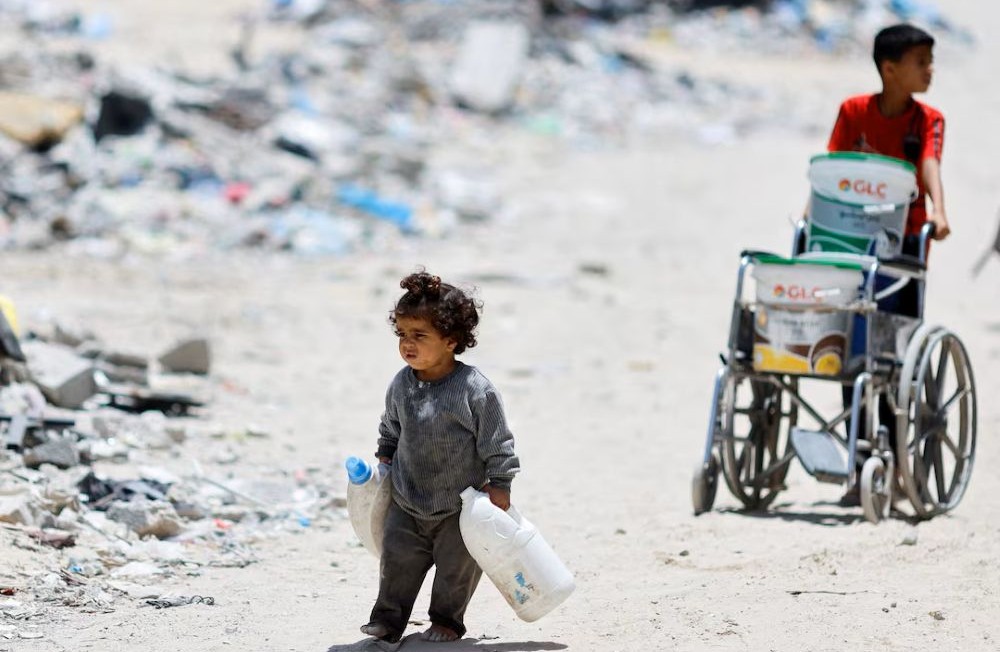 A Palestinian girl carries plastic bottles in which to collect water as she walks by houses that were destroyed by an Israeli strike, in Khan Younis, in the southern Gaza Strip, May 22, 2024. (Reuters)