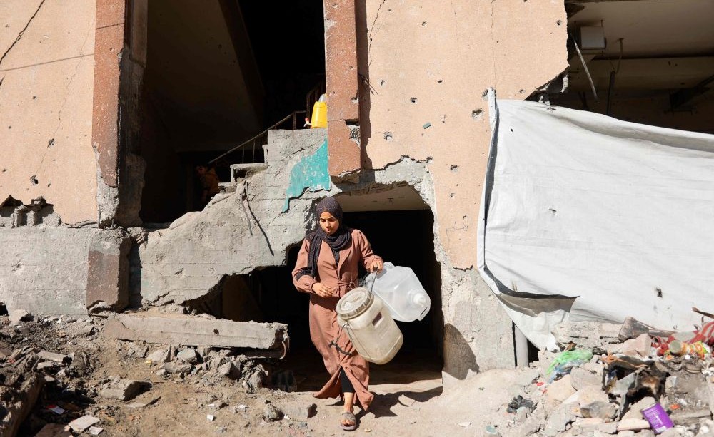 A displaced Palestinian woman, carrying water containers, exits a destroyed building, where families have set-up up shelters, Gaza City, Jan. 11, 2026. (AFP)
