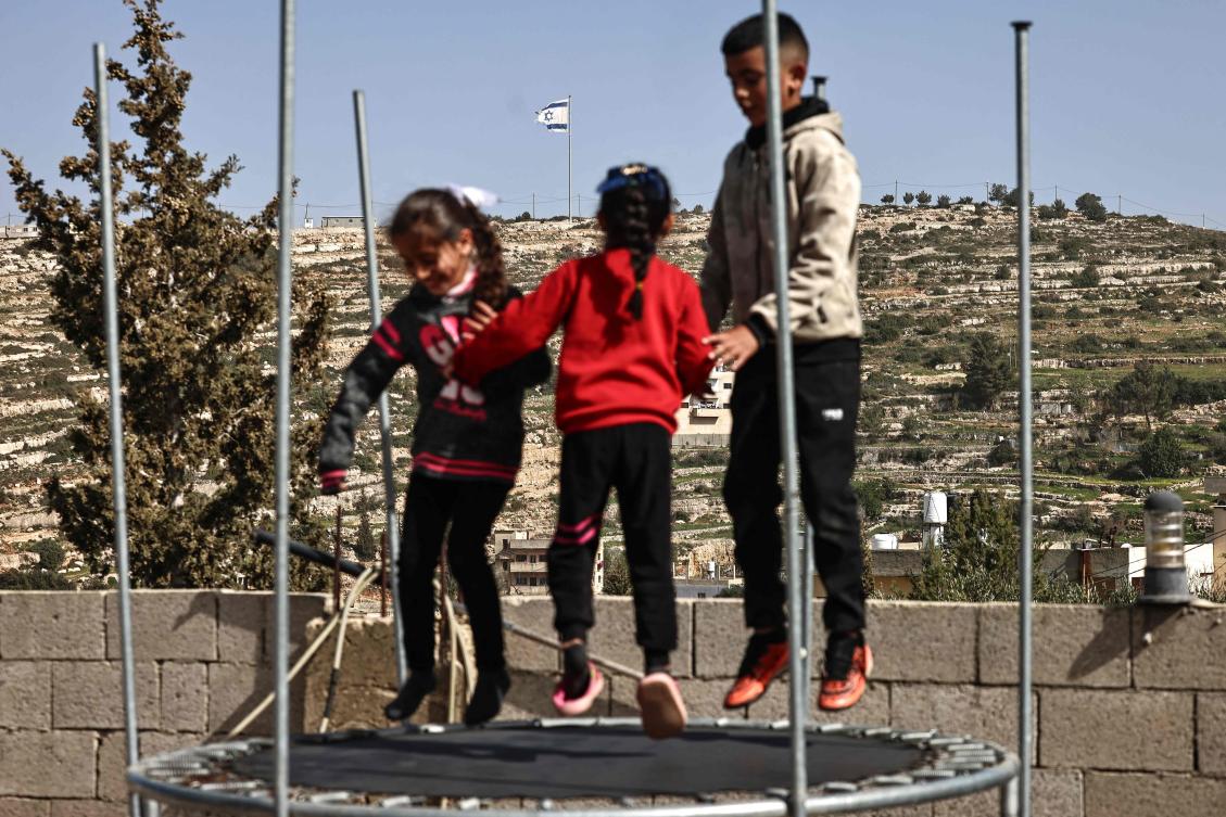 Palestinian children play on a trampoline in Umm Safa village, north of Ramallah, in the occupied West Bank, opposite an Israeli flag that was raised on a hilltop overlooking the village, Feb. 16, 2026. (AFP)