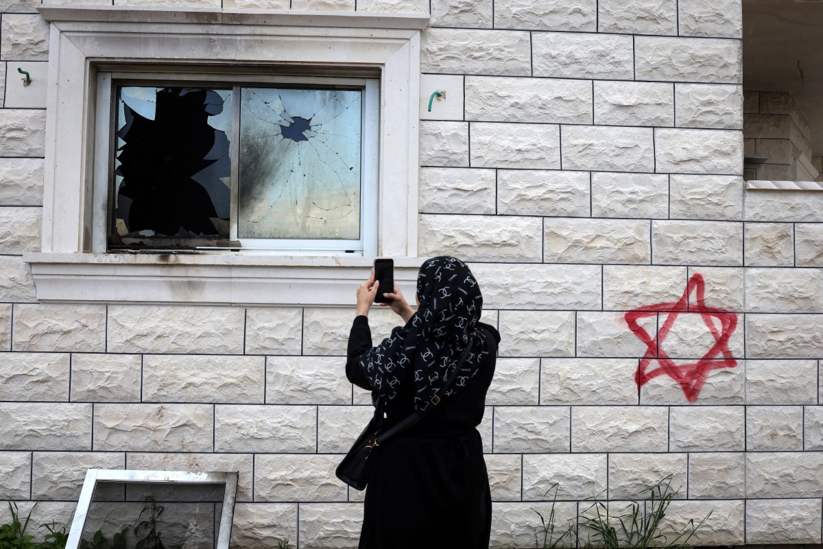 A Palestinian woman photographs a shattered window beside graffiti of a Jewish star of David sprayed on the wall of a damaged home following a reported attack by Israeli settlers in Silat al-Dhahr, in the occupied West Bank, March 22, 2026. (AFP)