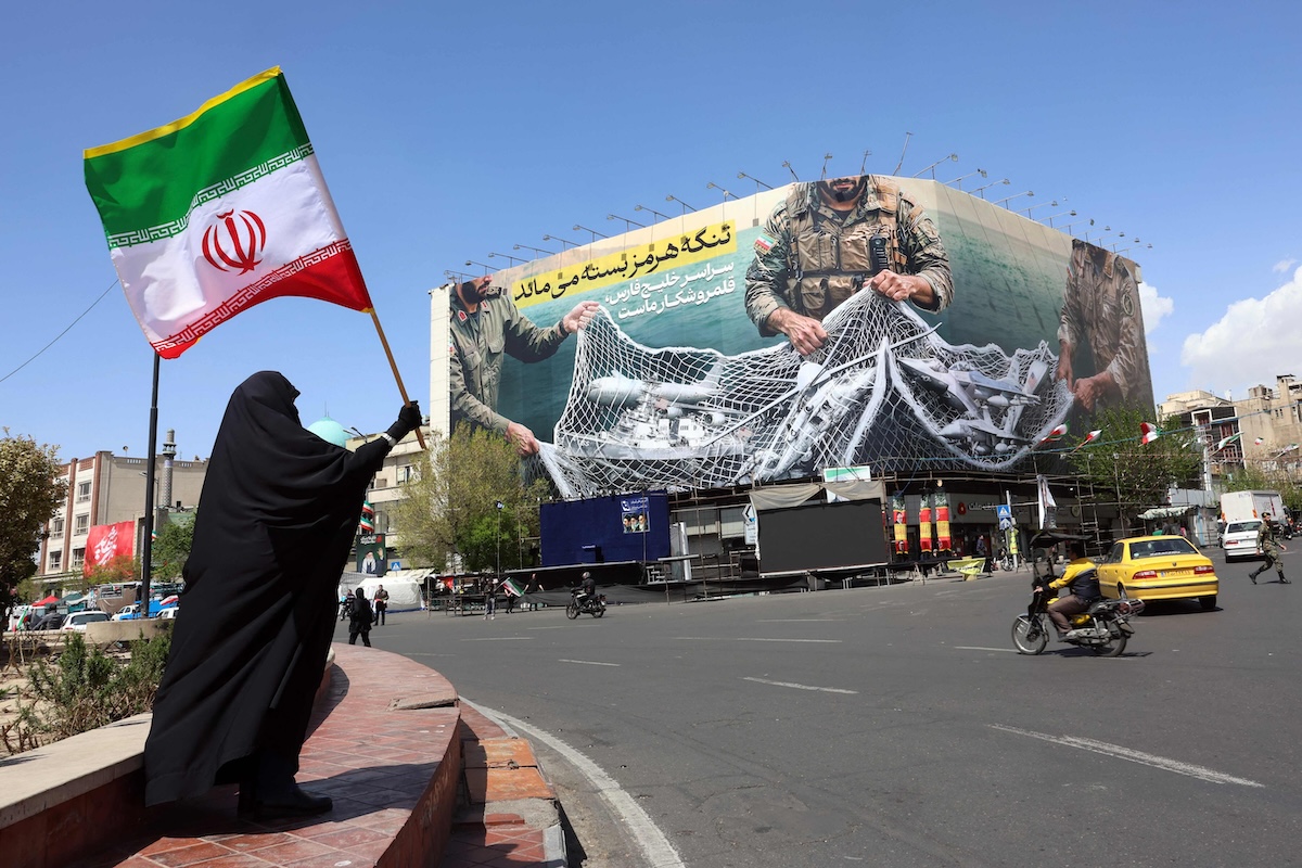 Above, a woman holds Iran’s national flag while standing near a billboard with a sentence reading ‘The Strait of Hormuz remains closed’ at the Enqelab Square in Tehran, on April 5, 2026. (AFP)