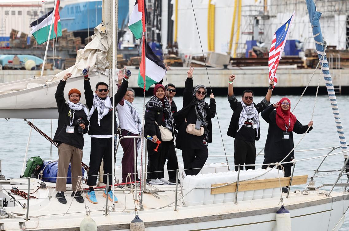 Pro-Palestinian activists wave onboard a boat of a new humanitarian flotilla bound for the Gaza Strip making a symbolic leave from Barcelona's Port Vell on April 12, 2026 as the departure of the flotilla has been postponed due to bad weather. A new flotilla of more than 80 boats and 1,000 international pro-Palestinian activists, who attempted to reach Gaza last year, is preparing for a new mission to the devastated territory from Barcelona. Israel's response to the Global Sumud Flotilla's first journey —intercepting the boats and arresting the activists— sparked international condemnation. (Photo by Josep LAGO / AFP)