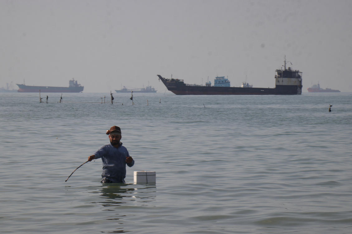 A man stands in the water as bulk carriers, cargo ships, and service vessels line the horizon in the Strait of Hormuz off Bandar Abbas, Iran, Monday, April 27, 2026. (AP)