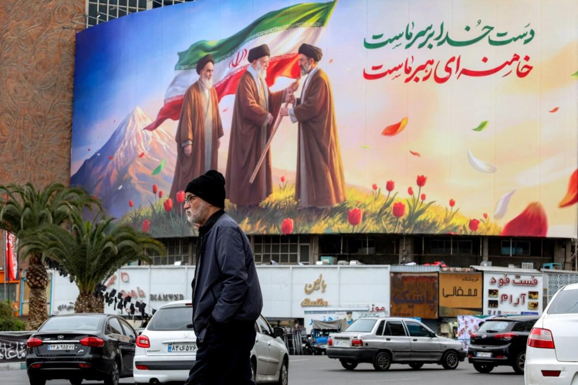 A man walks near a banner displayed at Valiasr Square in central Tehran on March 10, 2026, depicting Iran's late supreme leader Ayatollah Ruhollah Khomeini (L) watching as his successor the late Ayatollah Ali Khamenei (C) hands over a national flag to his son and new supreme leader Mojtaba Khamenei (R). (File/AFP)