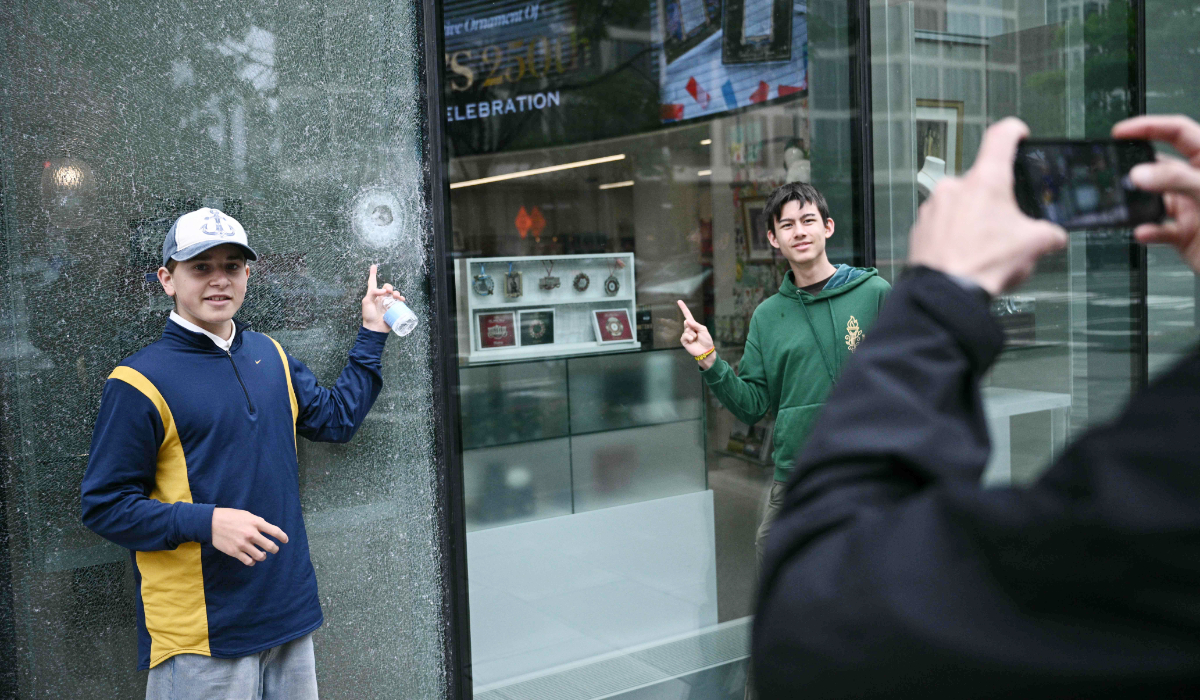 Two young men pose in front of the shattered window of a gift shop, which was struck by gunfire during a May 23 shooting, near the White House in Washington, DC, on May 24, 2026. (AFP)