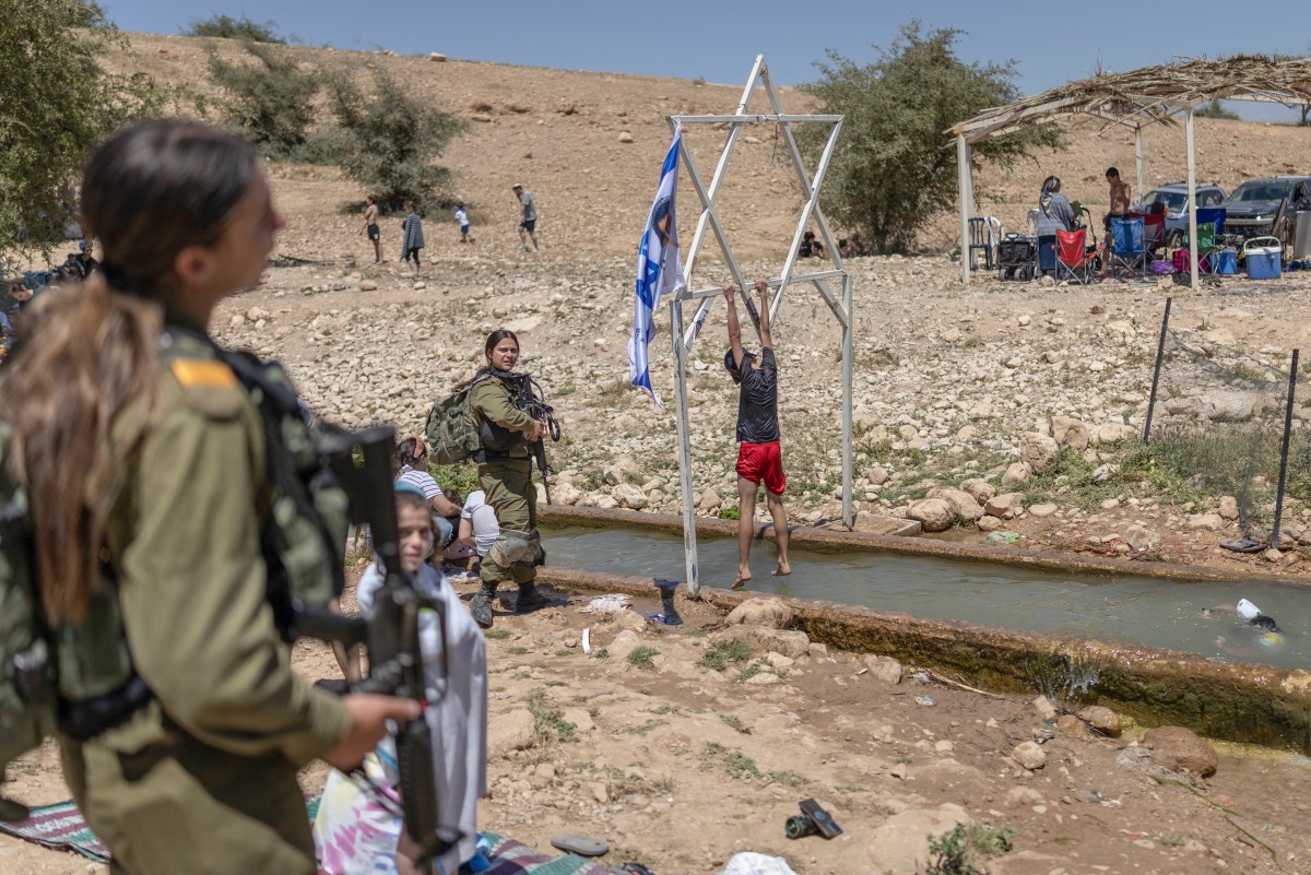 Armed Israeli women in military uniform stand with children as they enjoy the Palestinian water spring of Ein al-Auja as they celebrate the Israeli Independence Day at the Israeli-occupied West Bank village of Ras Ein al-Auja, whose Bedouin community was displaced by Israeli settlers earlier this year, Apr. 22, 2026. (Files/AFP)
