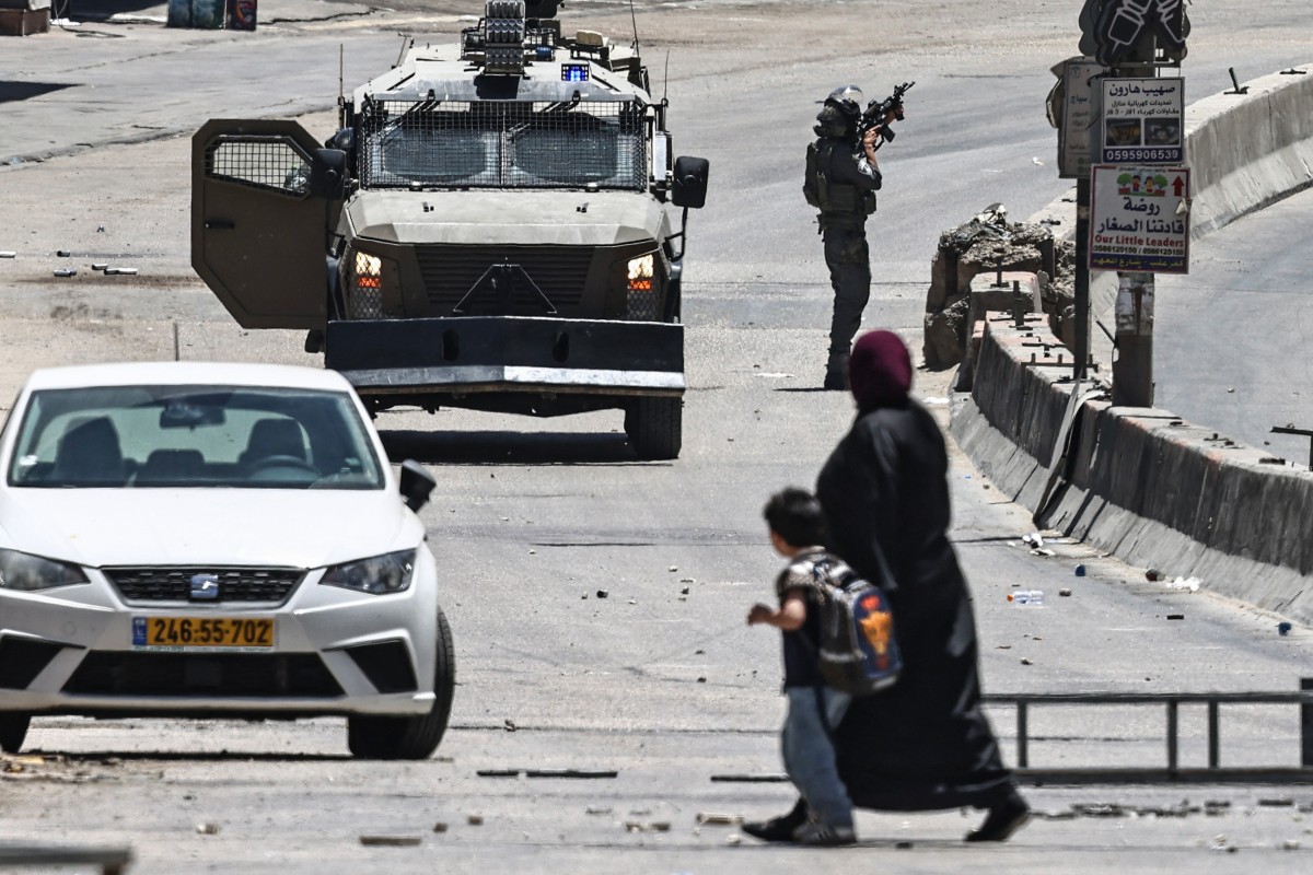 A woman and a child cross a street as Israeli security forces patrol during a military raid in the Qalandia refugee camp, south of Ramallah in the Israeli-occupied West Bank, May 11, 2026. (AFP)