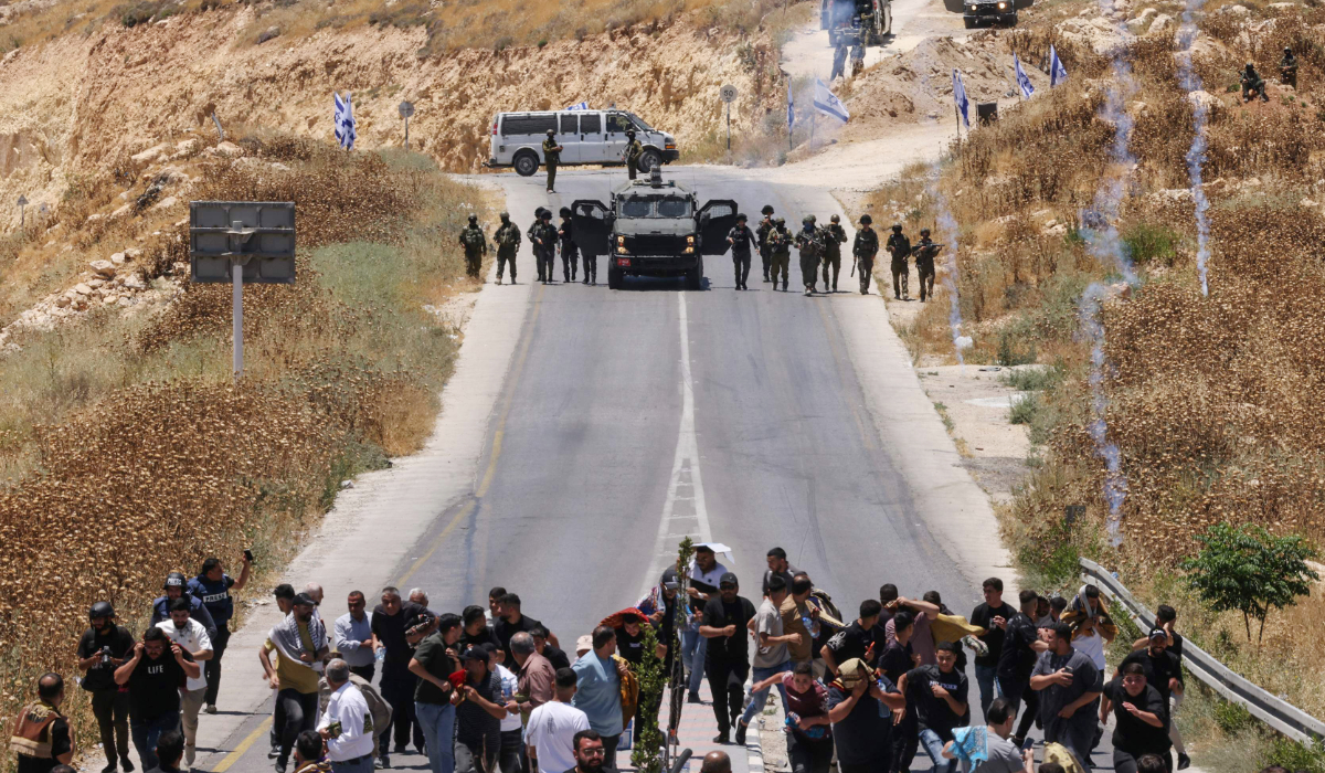 Protesters and local Palestinian landowners, who say some of their land was taken for the newly built Mount Tarousa settlement (background), run from tear gas fired by Israeli security forces blocking a road leading to the hilltop during a rally against the settlement near Dura in the Israeli-occupied West Bank on June 19, 2026. (AFP)