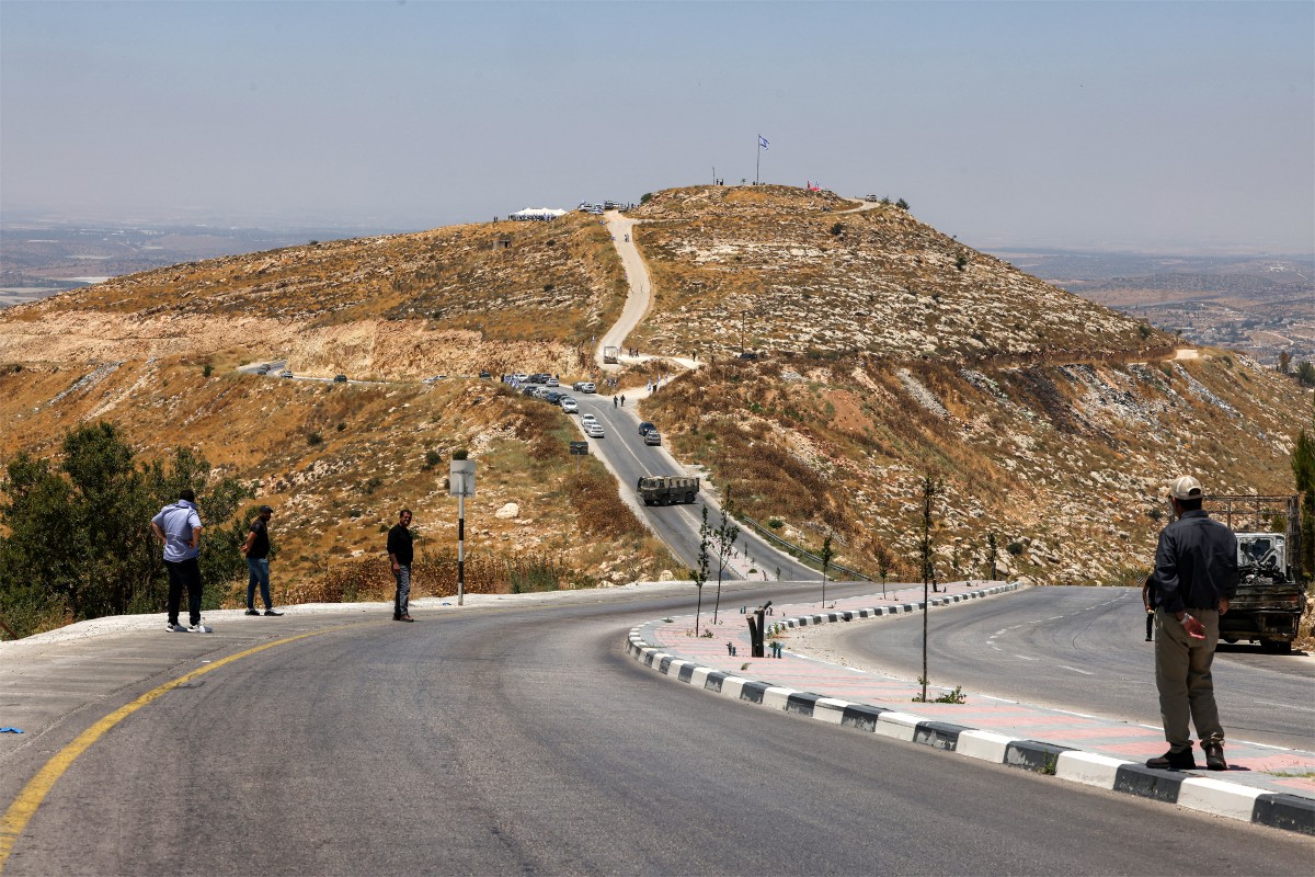 Palestinian men watch from a road as Israeli settlers gather in the background during a cornerstone-laying ceremony for a new settlement on Mount Tarousa, near the town of Dura, west of Hebron, Jun, 16, 2026. (AFP)