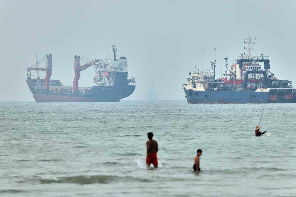 Children wade in the water with cargo ships at anchor in the background and a fisherman nearby, in the Strait of Hormuz off Bandar Abbas, Iran. (File/AP)