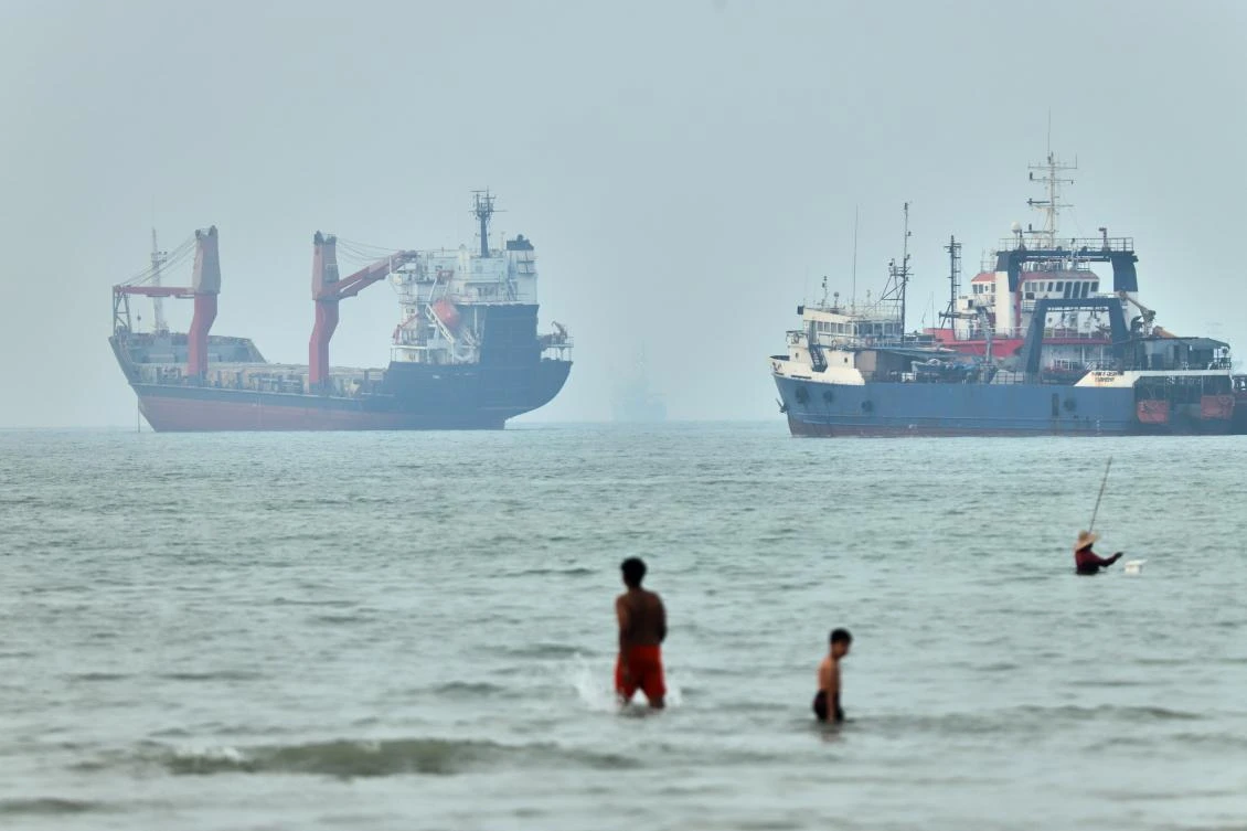 Children wade in the water with cargo ships at anchor in the background and a fisherman nearby, in the Strait of Hormuz off Bandar Abbas, Iran. (File/AP)