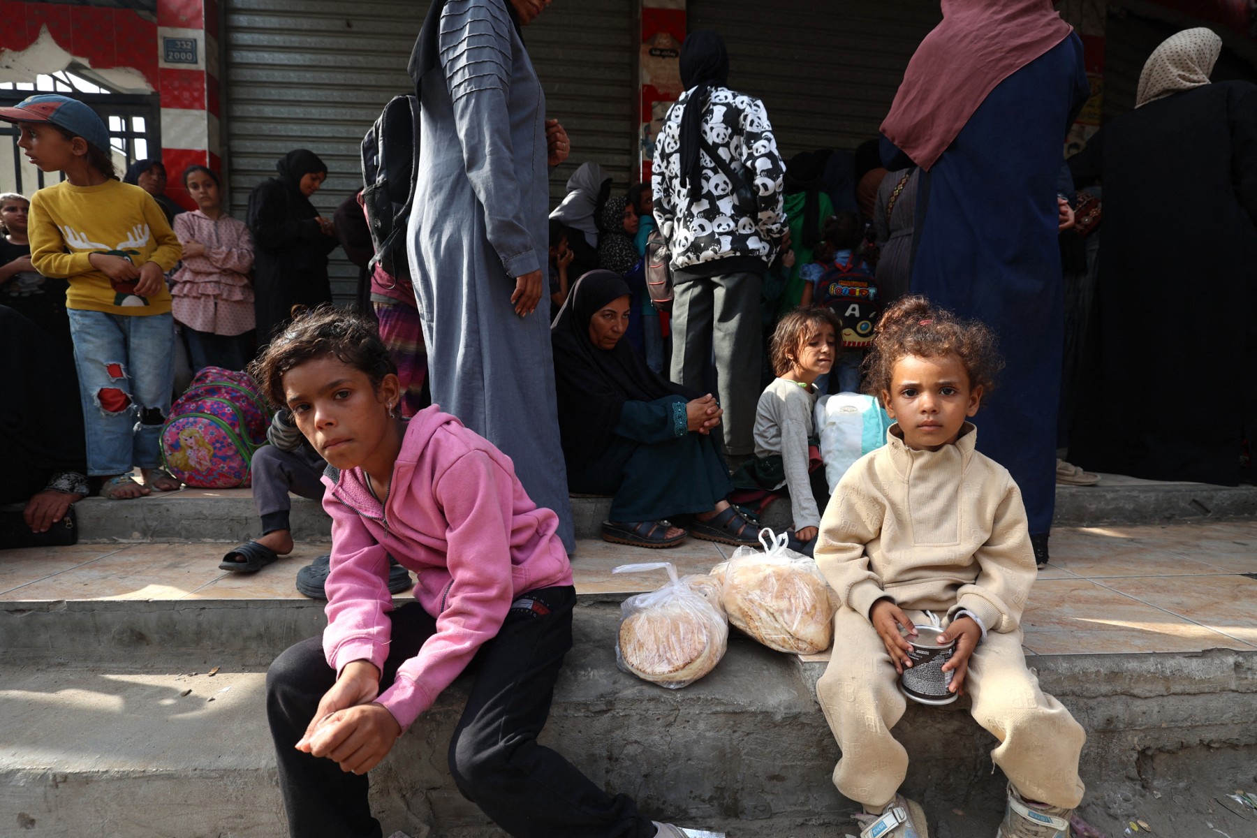 Palestinian children sit on the sidewalk after receiving bread portions from a charity bakery in the Nuseirat refugee camp, located in the central Gaza Strip. (File/AFP)