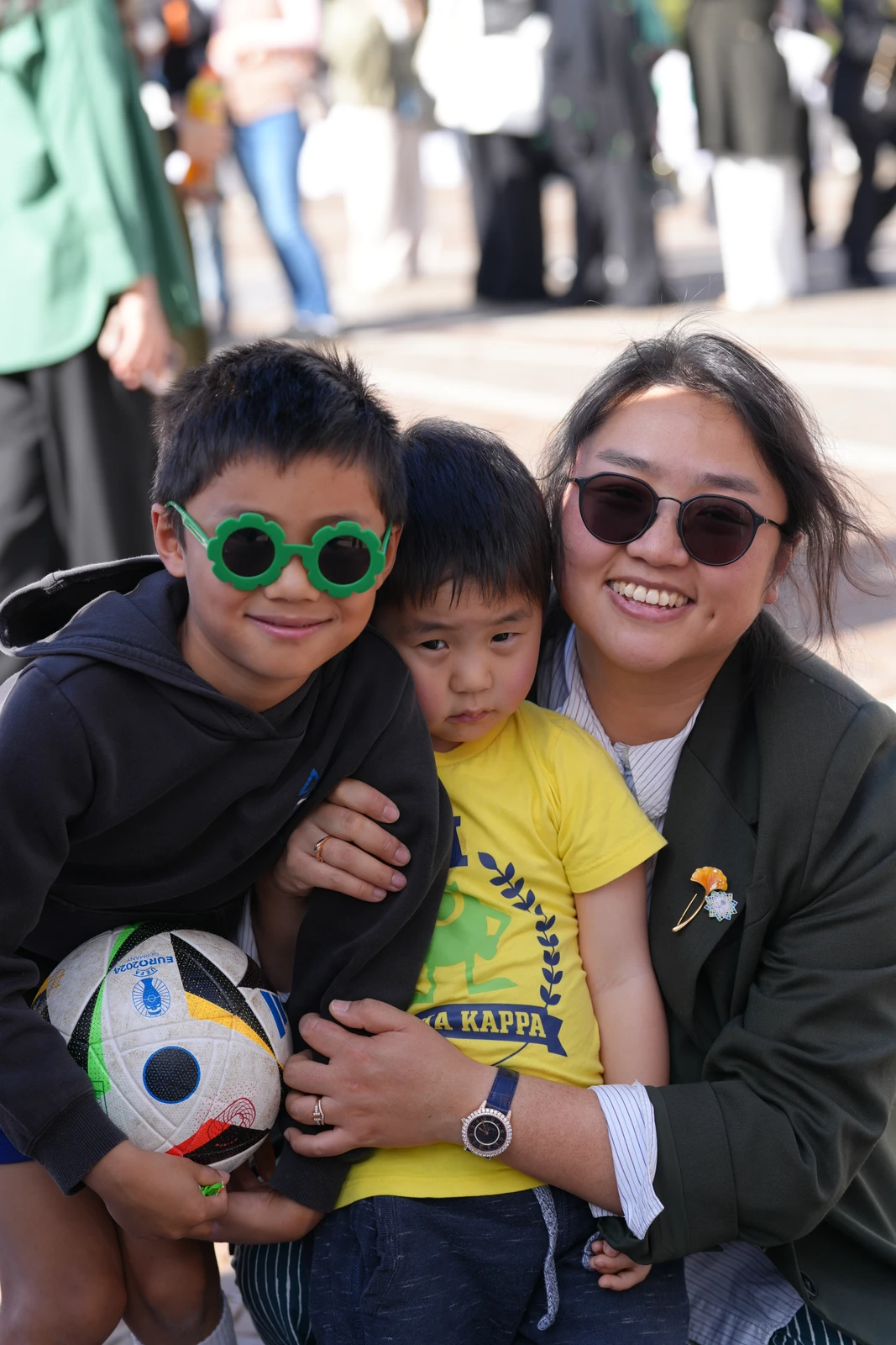 A smiling woman and two children pose together; one child wears green sunglasses and holds a soccer ball, and the other wears a yellow shirt.