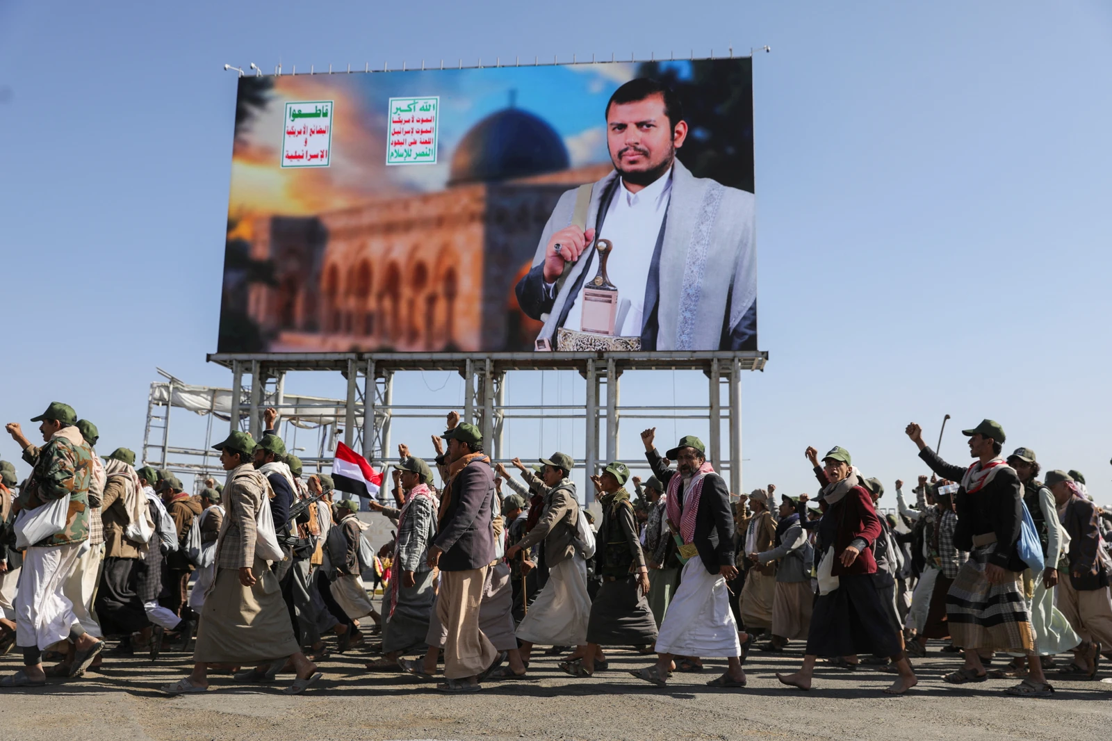 Houthi-mobilized fighters parade in Sanaa, Yemen, beneath a large billboard. The billboard features a portrait of a man, possibly Omar Souleyman or Brian Robiskie, against a backdrop showing the Dome of the Rock. The parading men, some in military attire and others in traditional Yemeni clothing like thobes and futahs, raise their fists in solidarity under a clear blue sky, with a Yemeni flag visible among them.