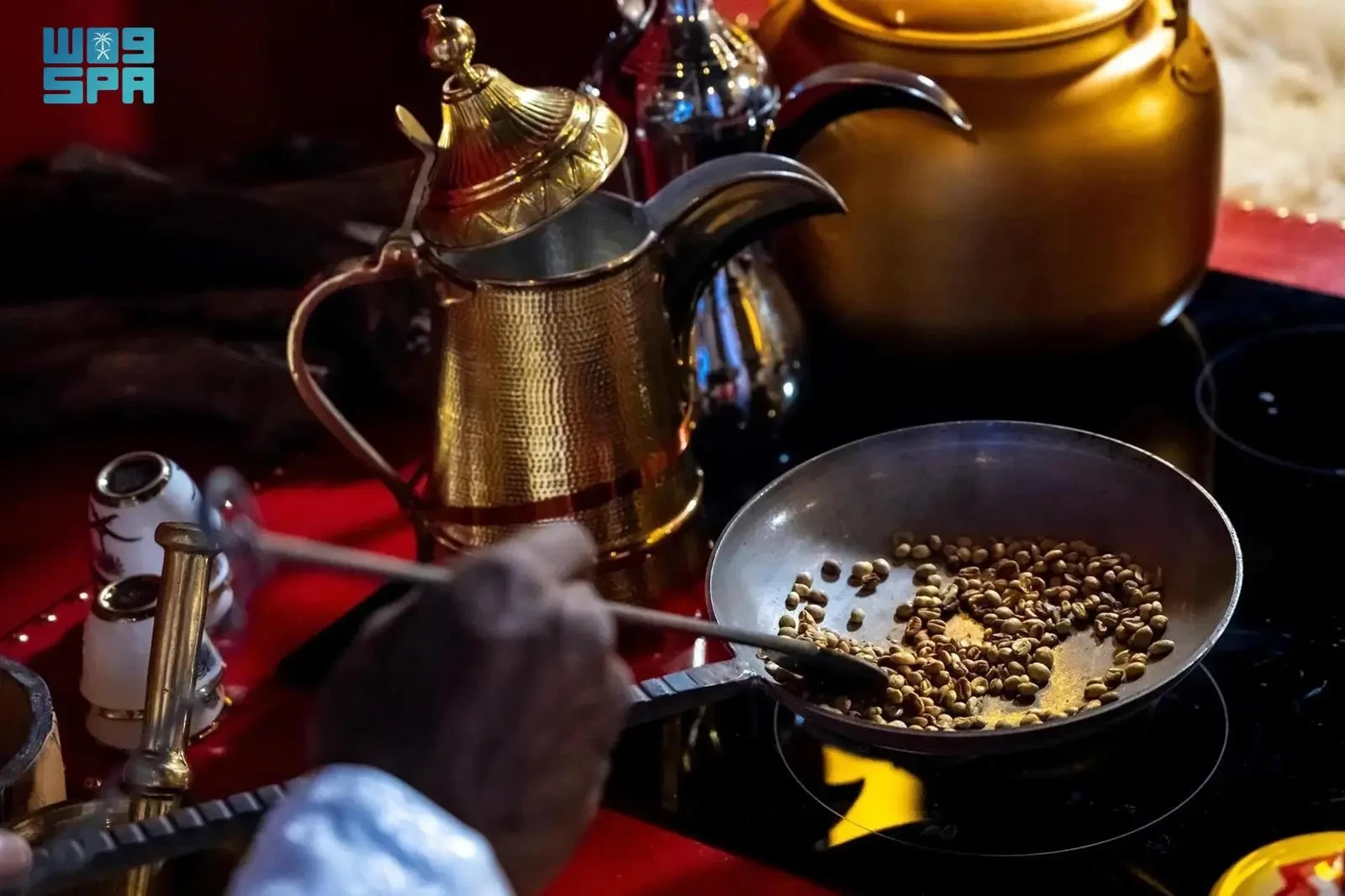Person roasting coffee beans in a frying pan with a golden pitcher nearby, set against a red background.