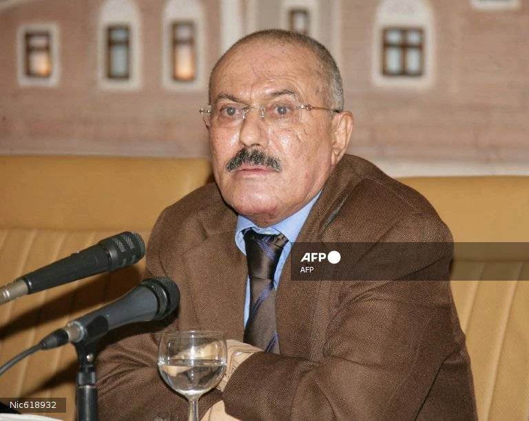 A middle-aged man with a mustache and glasses, wearing a brown suit and blue shirt with a tie, sits in front of two microphones and a glass of water, against a light brown background with decorative window-like shapes.