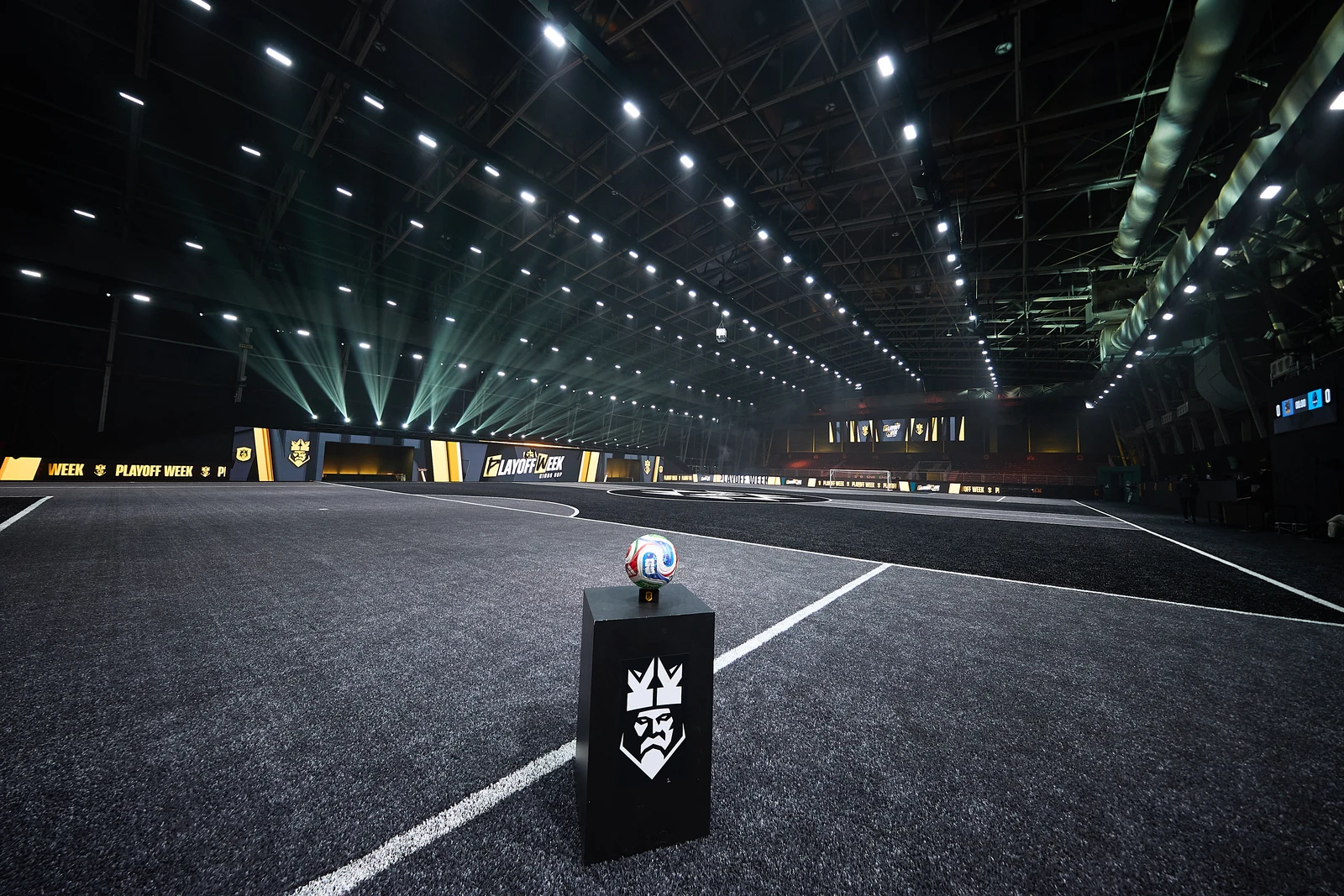 Interior of a large indoor sports arena with a lit black futsal field and a soccer ball on a pedestal.