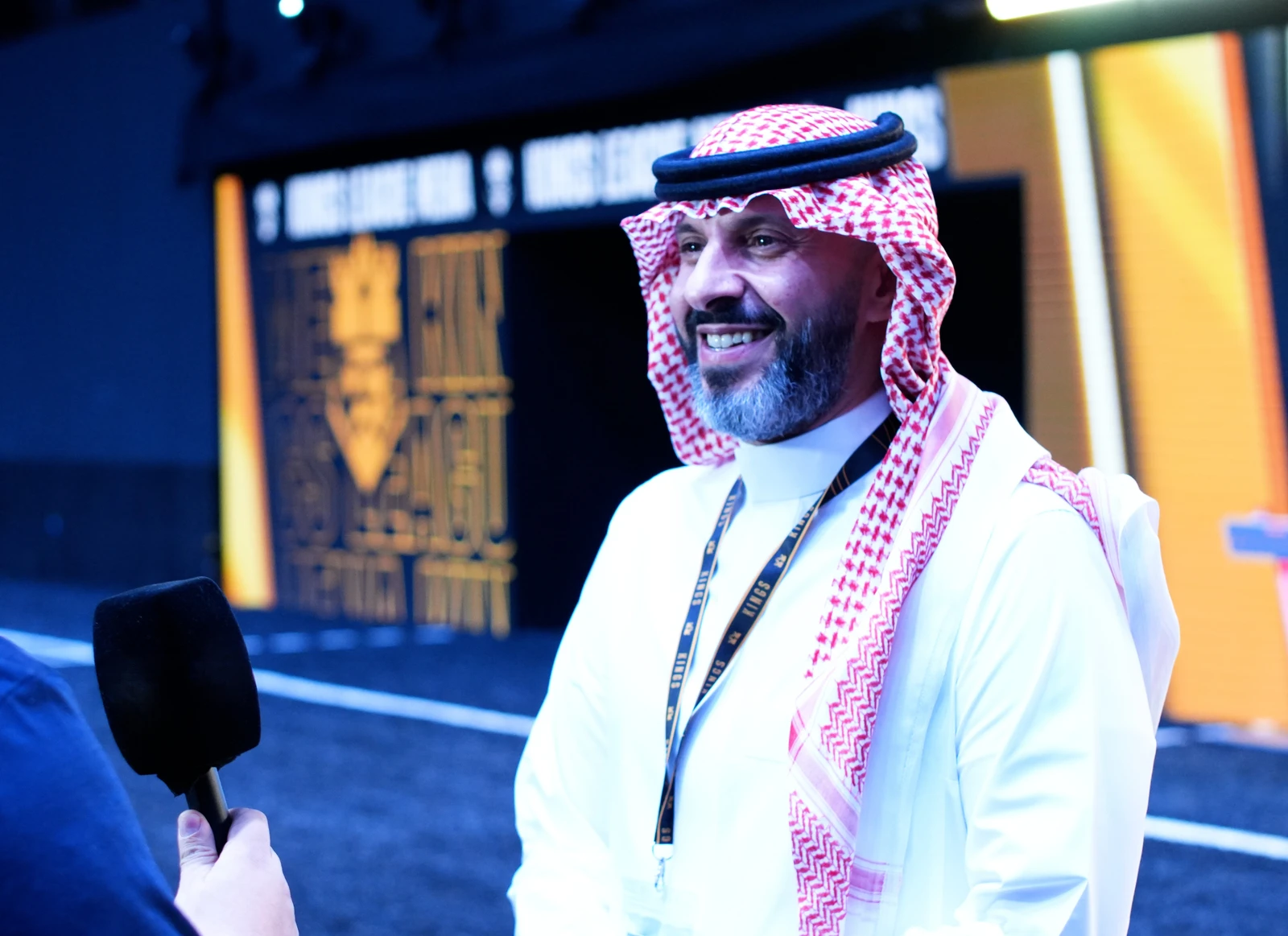 Aaidh ibn Abdullah al-Qarni being interviewed at an event, wearing traditional Saudi attire and smiling with a stage background.