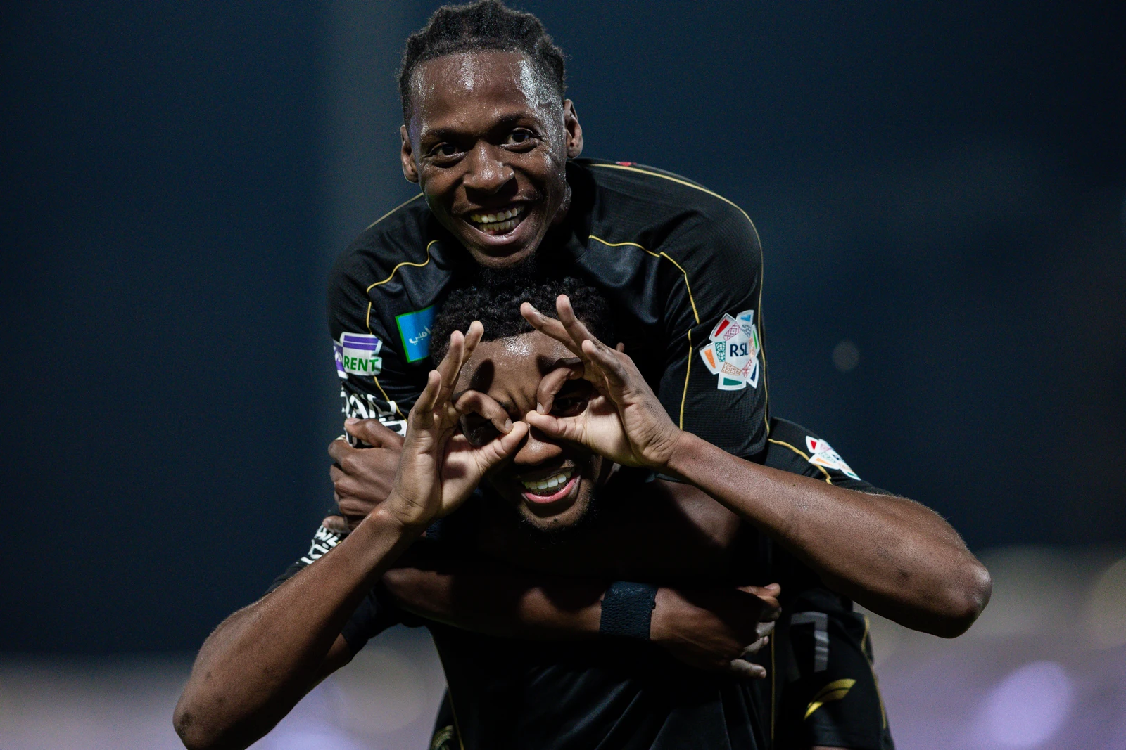 Two male soccer players celebrate a goal or victory on the field at night. The player on top has his arms around the neck of the player below him. Both are smiling widely, and the player on the bottom is making a funny 'goggles' gesture with his hands over his eyes.