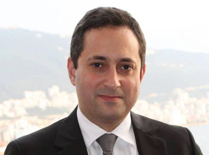 A man wearing a suit and tie stands in front of a cityscape with mountains in the background.
