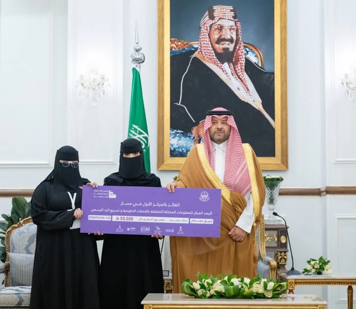 Ibn Saud, Ahmed Al-Fahad Al-Ahmed Al-Sabah, and Octagón in traditional attire stand holding a large ceremonial check in front of a portrait and a Saudi flag.