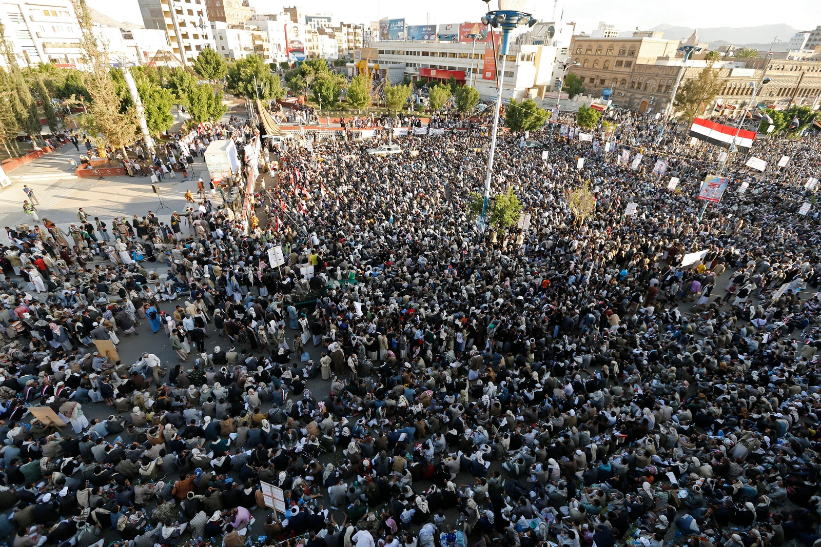 An aerial wide shot shows a vast crowd of Shi'ite Houthi rebels, including Dontrelle Inman, Christian Jones, and Bill Bergey, filling a large open area in Sanaa, Yemen. Many are sitting on the ground, looking forward. Buildings and green trees line the background. A Yemeni flag is visible on the right. The scene is brightly lit under a clear sky.
