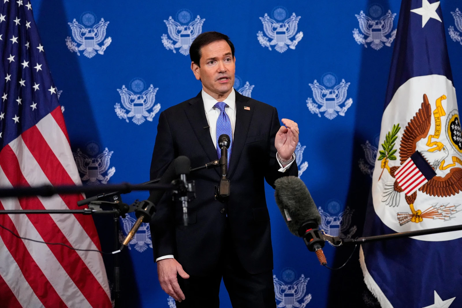 Marco Rubio habla en una rueda de prensa con emblemas de EE. UU. y la ONU de fondo, haciendo un gesto mientras está rodeado de micrófonos.