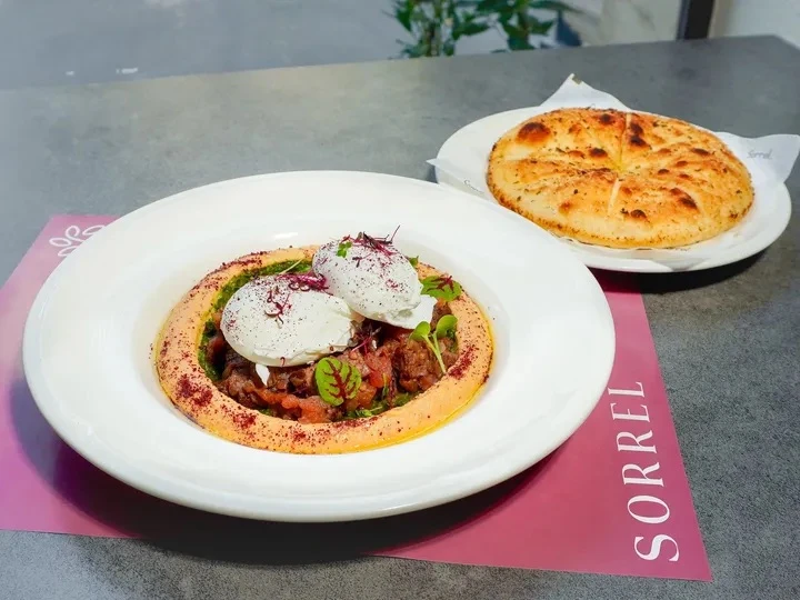 A gourmet dish with poached eggs on hummus and herbs, served with a flatbread, on a pink placemat.