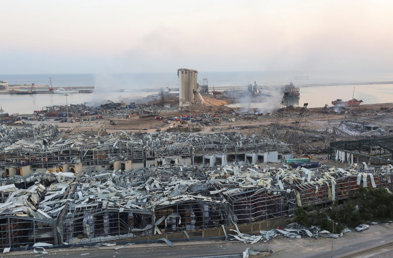Wreckage and smoke at an industrial site near a harbor, showing extensive damage and collapsed buildings.