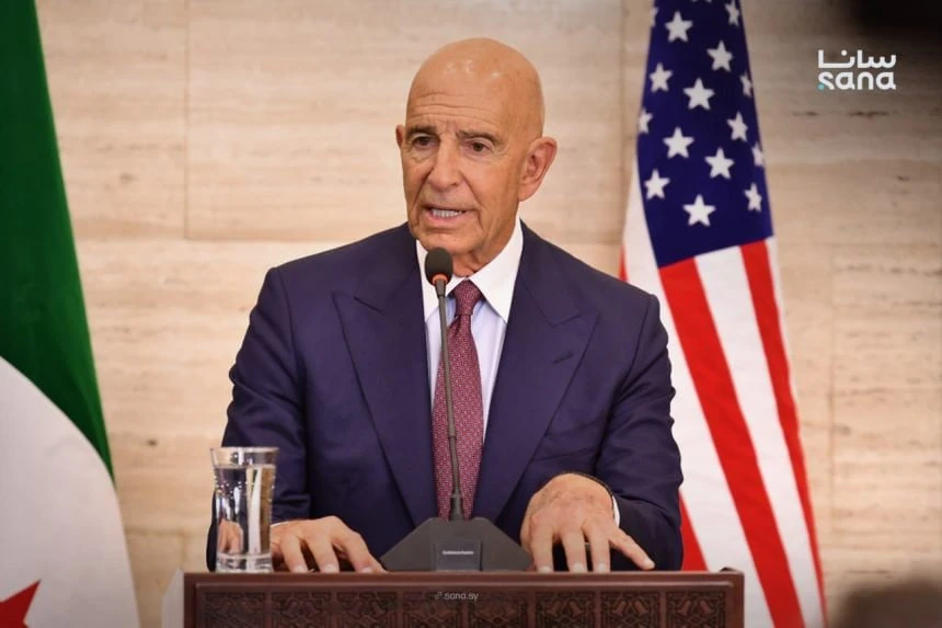 Thomas J. Barrack, Jr., a bald man in a dark suit and red patterned tie, speaks intently into a microphone at a wooden podium. A glass of water is to his left. Behind him are the American flag on the right and a green and white flag, likely the Algerian flag, on the left.