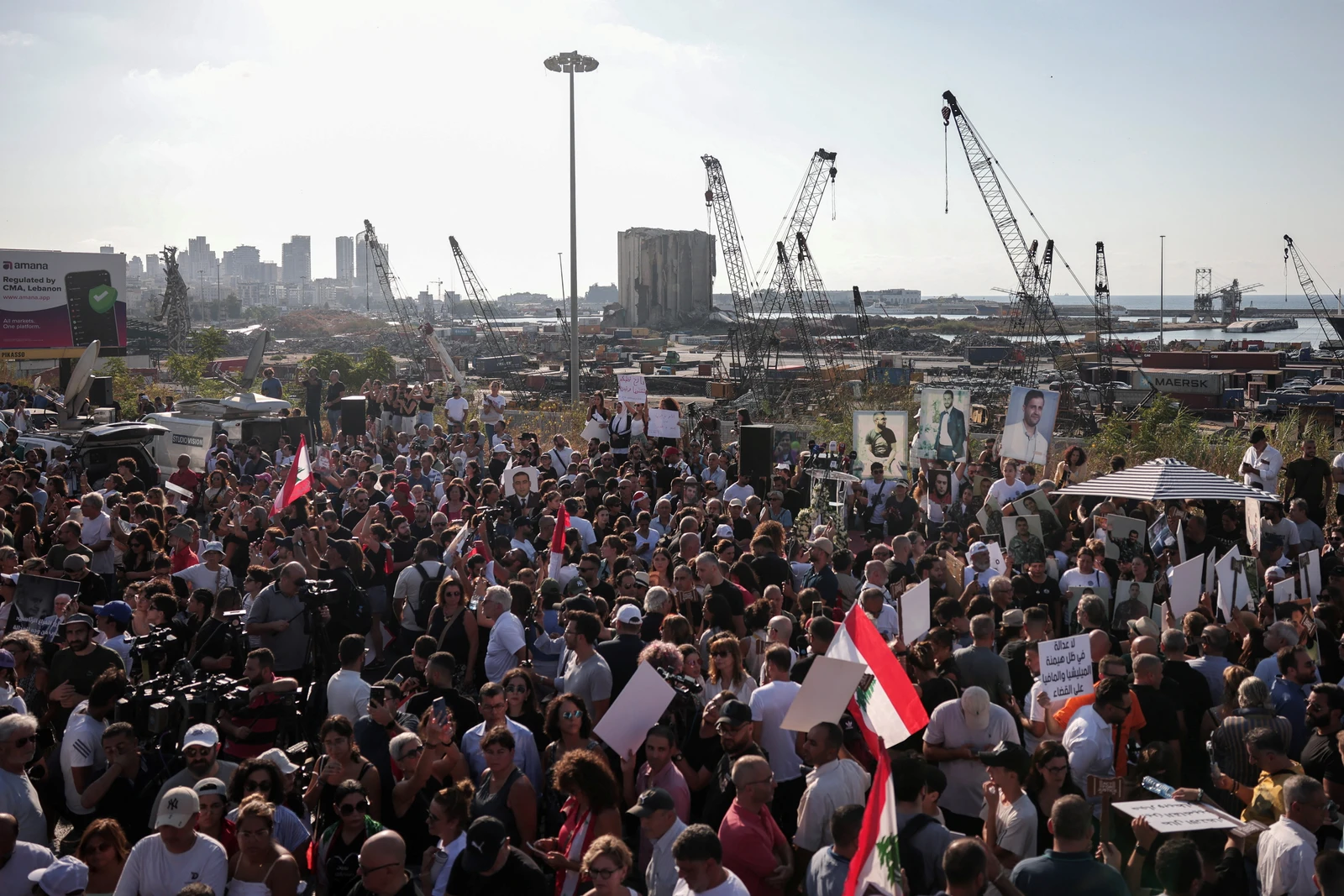 A large crowd protests near a port with cranes and city buildings in the background, holding flags and signs.