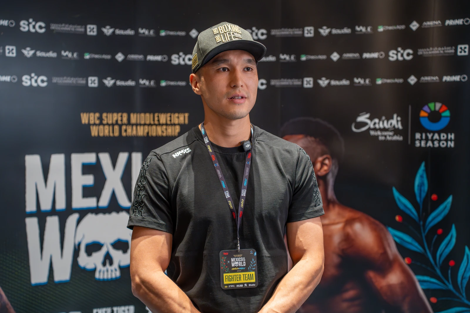 A man, possibly a boxer or athlete, wearing a black t-shirt and a baseball cap that reads "NO BOXING NO LIFE" is captured from the chest up, looking intently to the right. He has a serious expression, and a lanyard with an event pass hangs around his neck. The background is a dark banner with white and gold text, including sponsor logos and event titles.