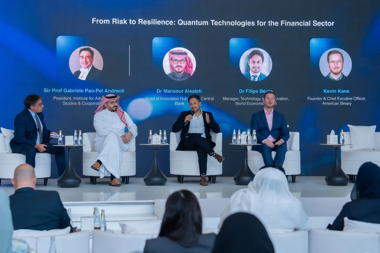 Four speakers seated on stage discussing quantum technologies for the financial sector with a digital backdrop displaying their names and portraits.