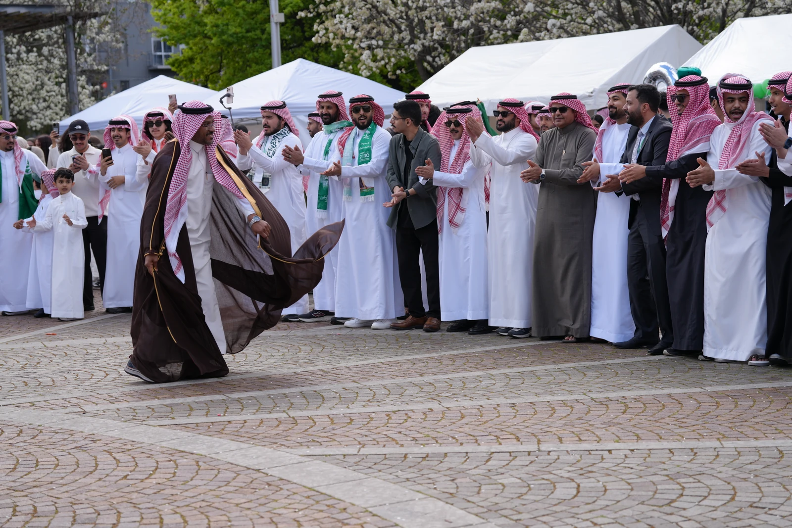 A man in traditional attire is dancing outdoors, with a group of men in similar attire clapping and cheering around him.