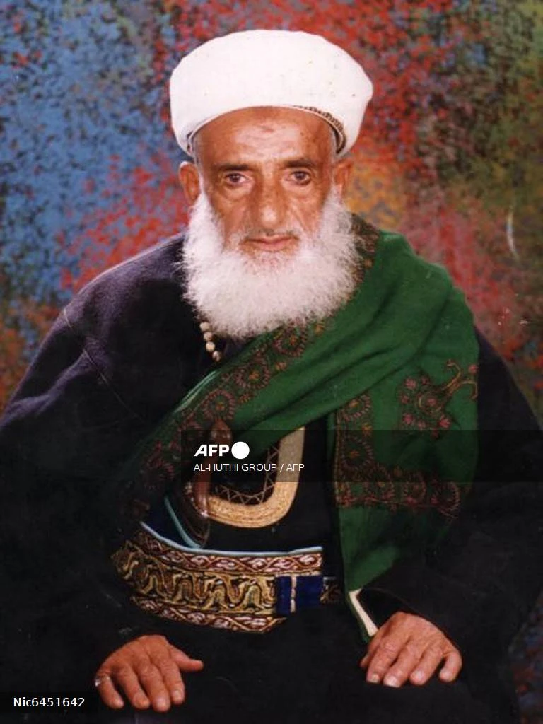 A close-up portrait of an elderly man with a long white beard and a white turban, wearing dark clothes, an embroidered belt, and a green scarf, looking seriously at the camera.