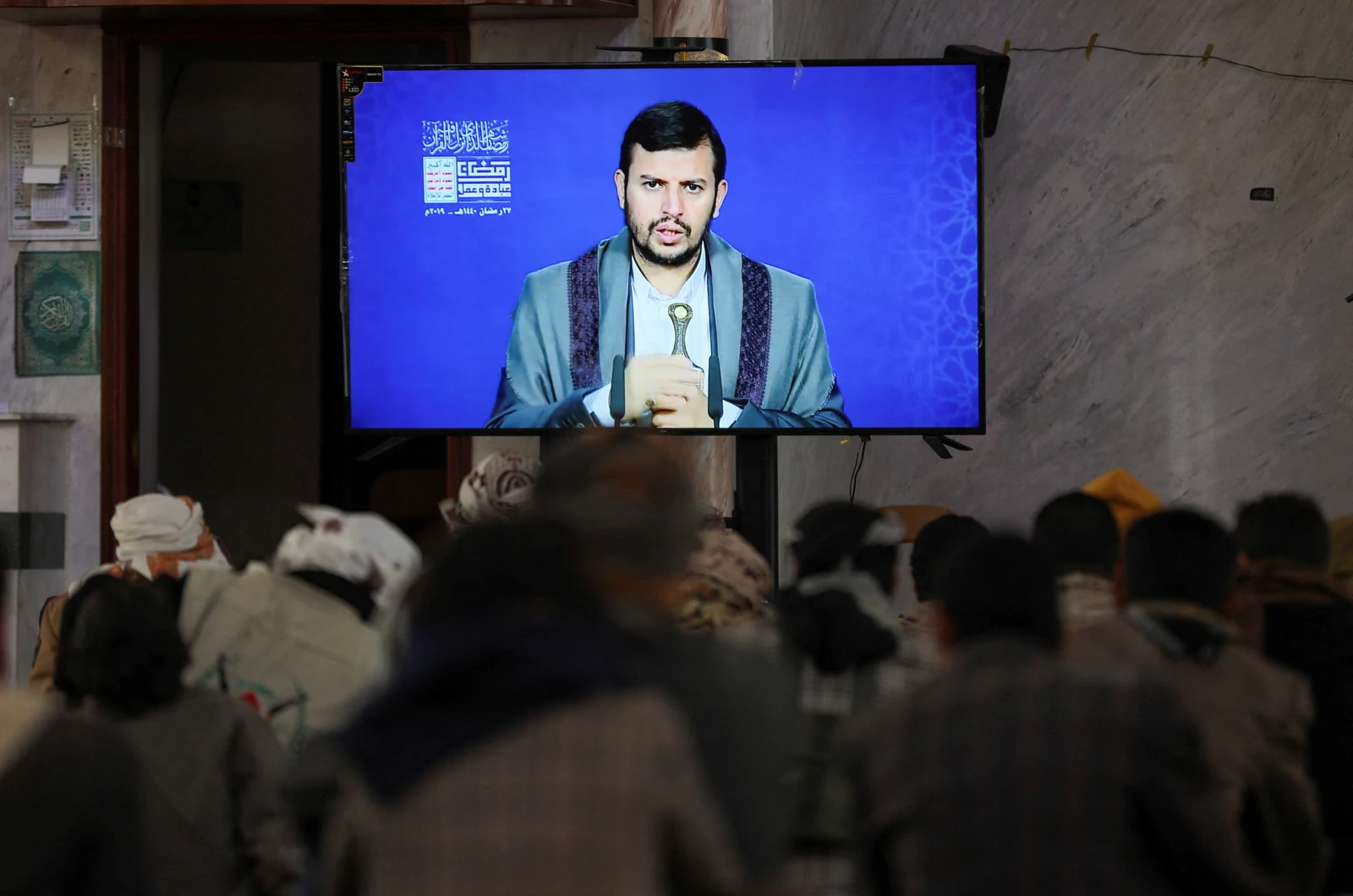 A group of Houthi fighters, seen from behind, are seated in an indoor room, watching a large television screen. The screen shows Houthi leader Abdul-Malik al-Houthi delivering a lecture. The fighters' heads and shoulders are visible, indicating an audience.