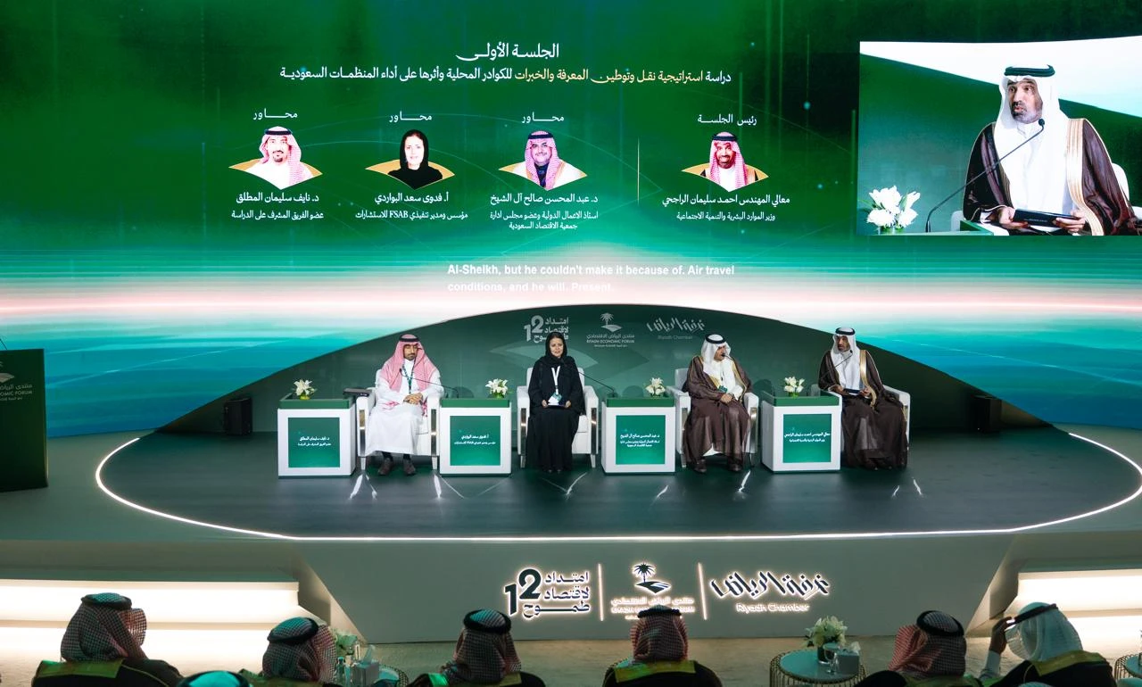 A wide shot of a modern conference stage. On the stage, Khalifa bin Zayed Al Nahyan, Claire Sugden, and three other individuals are seated in chairs, facing the audience. Behind them is a large screen showing speaker profiles and conference details, including Khalifa bin Zayed Al Nahyan in a close-up image. The stage is lit with green and white lights, and the overall ambiance is professional and organized.