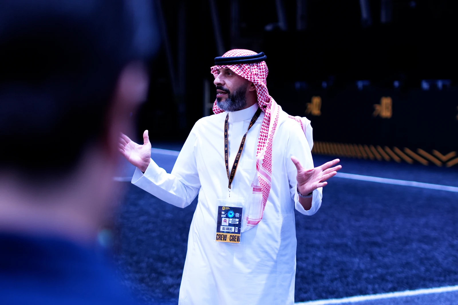 A man in traditional Saudi clothing, with a keffiyeh and a lanyard, gesturing as he speaks in an indoor setting.
