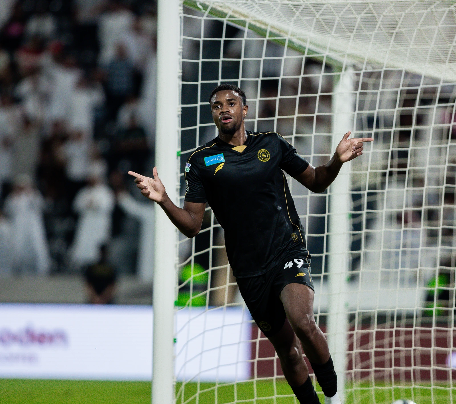 A male soccer player in a black and gold uniform with the number 49 on his shorts stands by the goal, arms wide, pointing to the sides in celebration. The white netting of the goal is prominent, and the background is softly blurred with stadium lights.