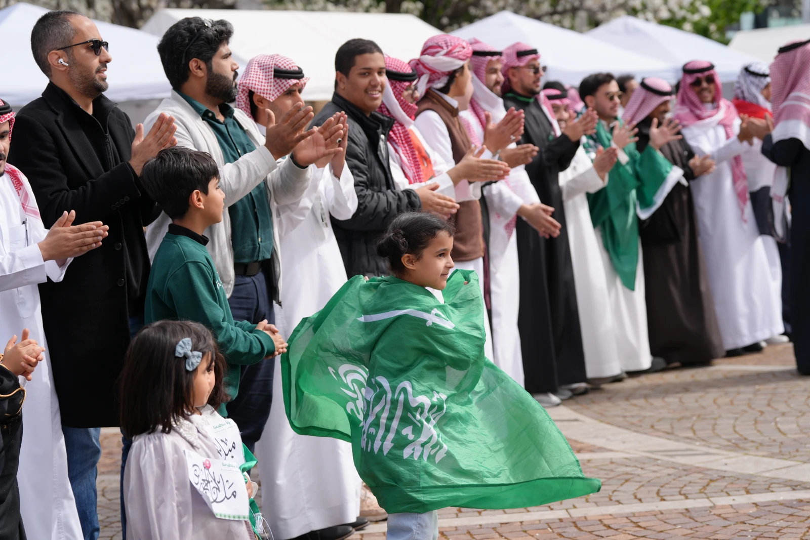 A diverse group of people, including children draped in green flags, participating in an outdoor celebration with traditional attire and a festive mood.