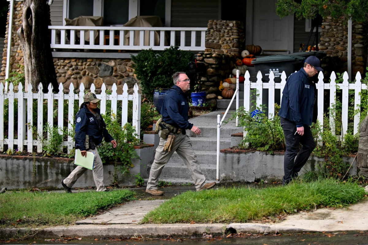 Three Homeland Security Investigations agents walk on a sidewalk. Lyle Sendlein, in a dark blue jacket and khaki pants, walks forward, looking right. To his left, another agent in a dark jacket and camouflage hat carries a light green folder. To his right, a third agent in a dark jacket and cap walks with a small white object in his hand. A white picket fence runs parallel to the sidewalk, with a house adorned with fall pumpkins in the background.