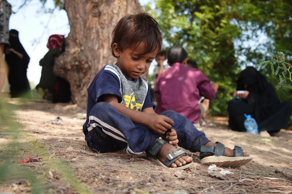 A displaced Yemeni child sits under a tree as families gather to receive aid at the Al-Fallahine displacement camp in Dar Saad district, Aden, Yemen. (File/AP)