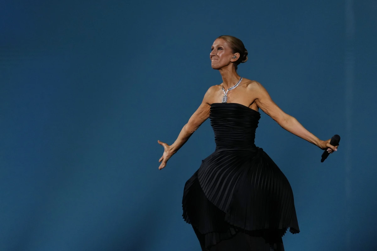 A singer in a black evening gown stands on stage with a microphone, against a blue background, striking a dramatic pose.