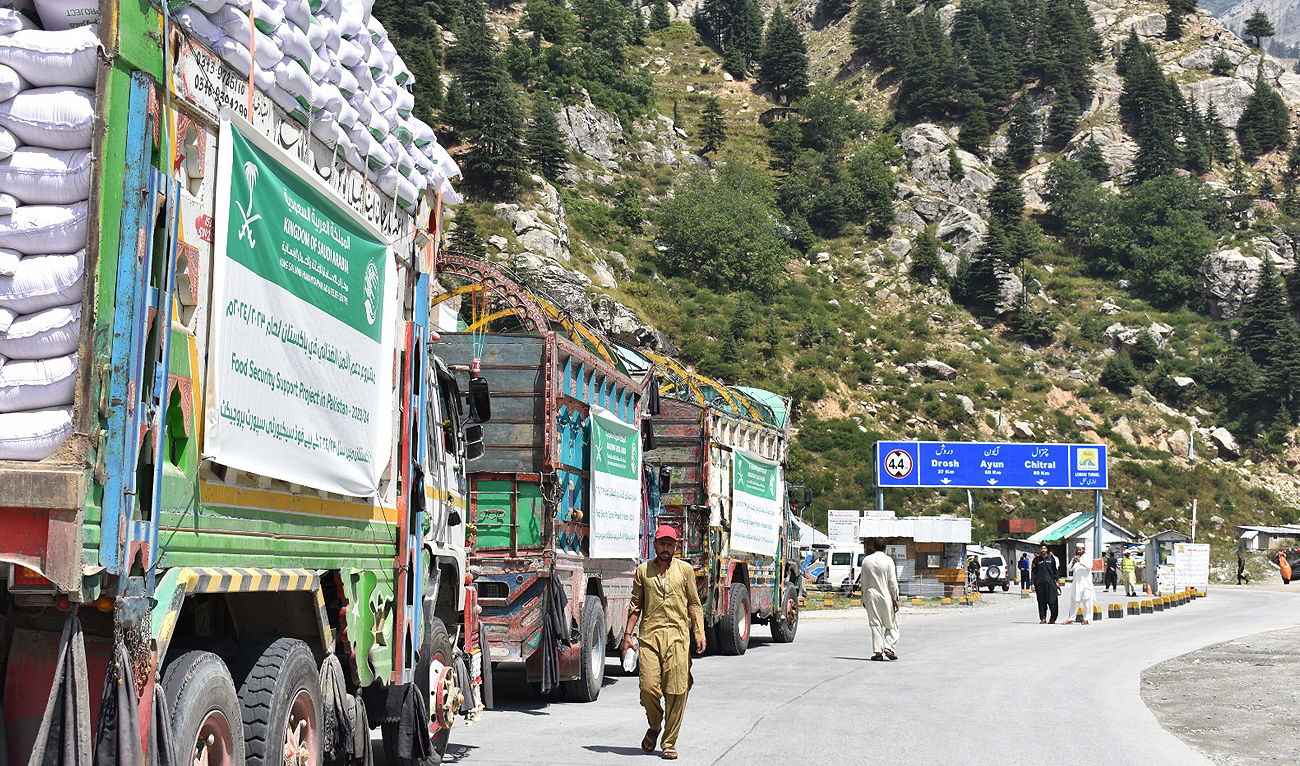 People walk past trucks loaded with food packages from King Salman Humanitarian Aid and Relief Centre (KSrelief) for flood victims in Chitral and Dir districts of northern Pakistan on September 10, 2023. (Photo courtesy: KSrelief)